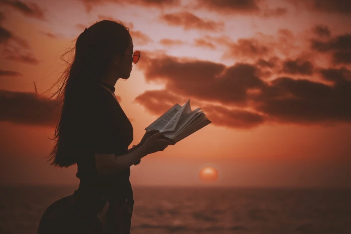 woman holding book while looking at body of water during golden hour Reading with sunset 2k 4k 5k