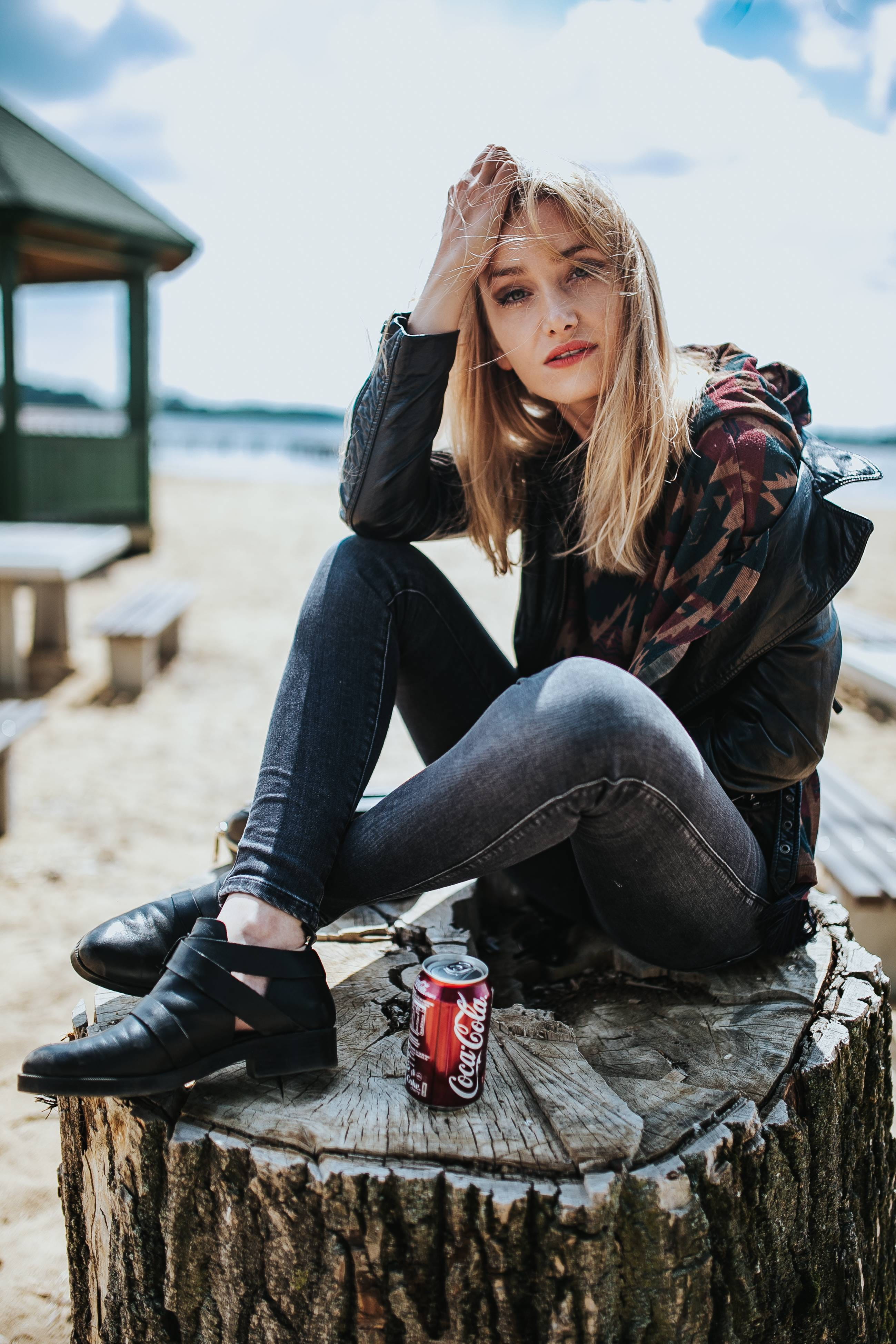 Beautiful blonde woman relaxing with a can of coke on tree stump by the beach 2k