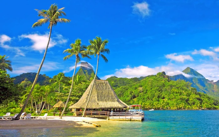 Tropical scenery coast palm trees huts bungalows mountains blue sky coconut trees; white wooden lounge chairs; brown tiki hut 2k