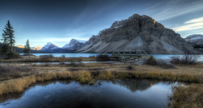 lake between grass field near sea and mountains under gray sky bow 2k