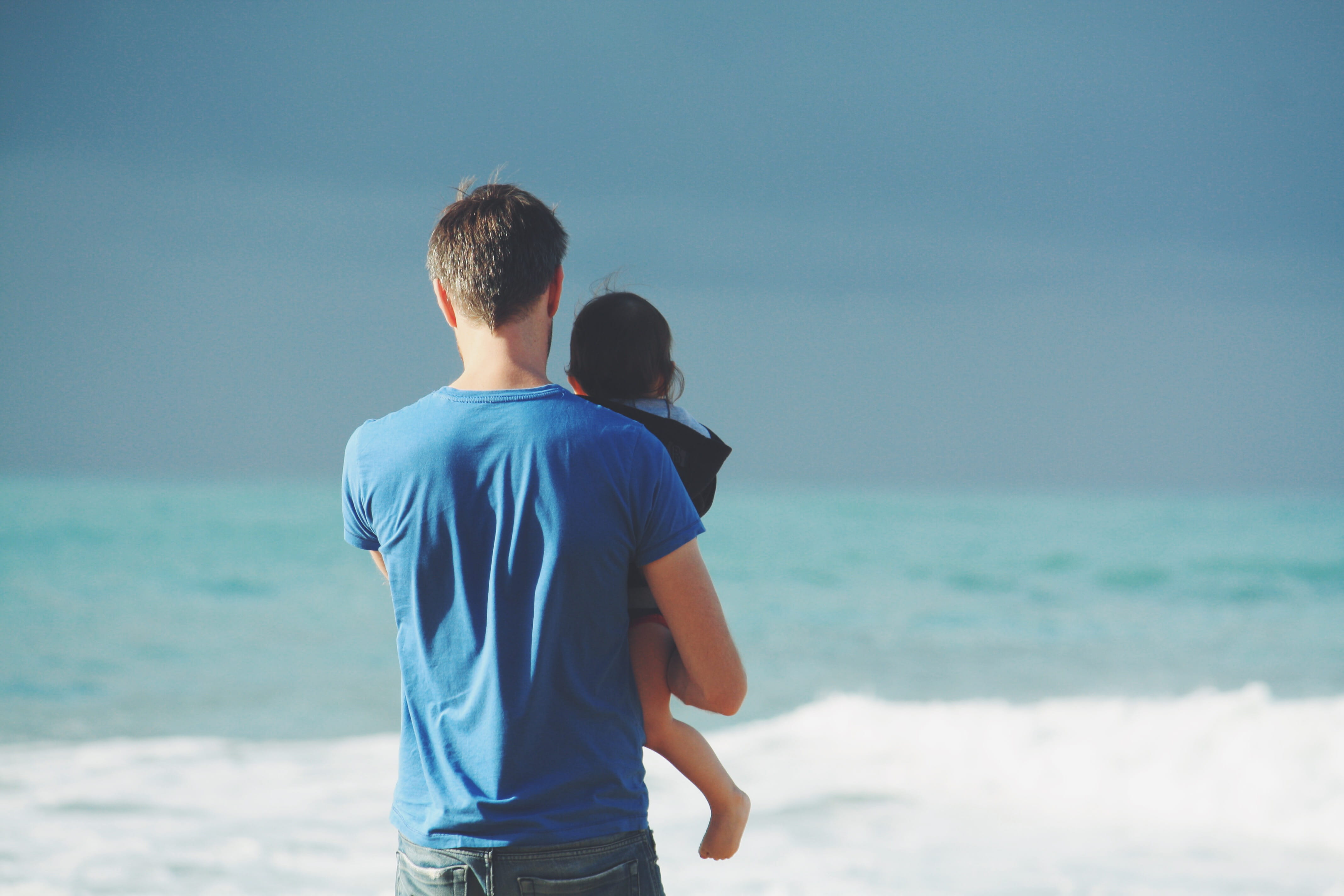 man wears blue crew neck t shirt holding toddler black hooded jacket near ocean under sky at daytime carrying body of water 2k 4k