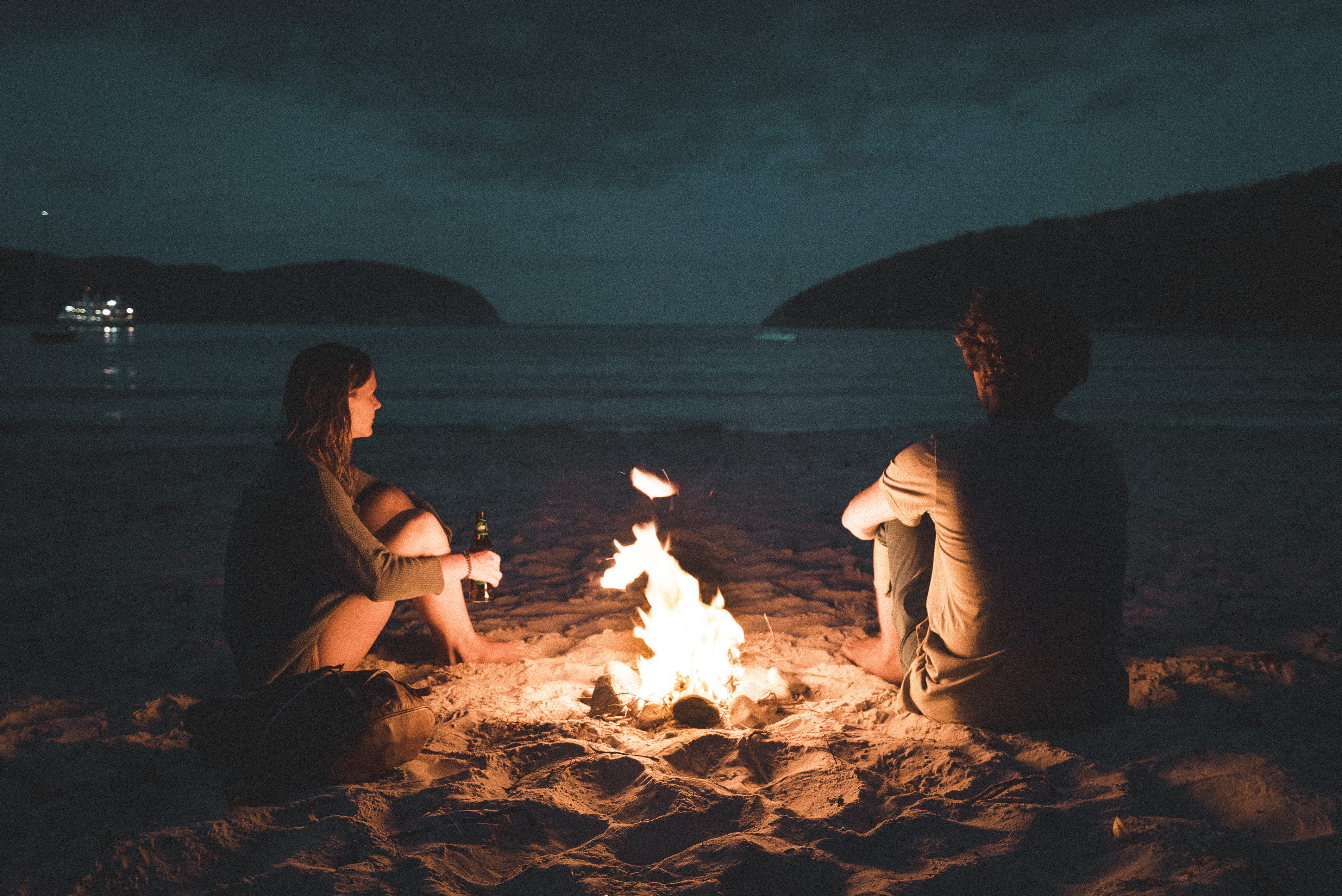 man and woman with bone fire sitting on seashore bonfire between near sea during nighttime 2k 4k